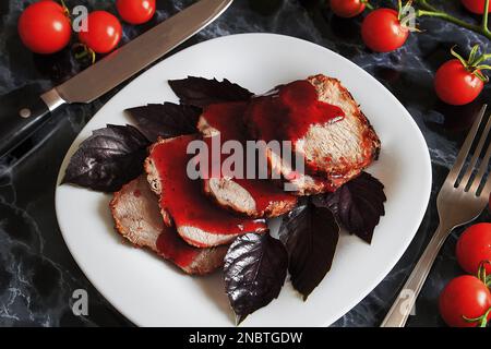 Saftiges Steak, gut gebratenes Schweinefleisch mit roter Sauce und Basilikumblättern auf einem weißen Teller über einem schwarzen Marmortisch. Kirschtomaten und Basilikum-Zweige auf einer mär Stockfoto