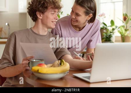 Ein junges, wunderschönes weißes Paar, das passende T-Shirts trägt, ein gesundes Frühstück hat, lacht und sieht sich an, sitzt am Tisch. Stockfoto