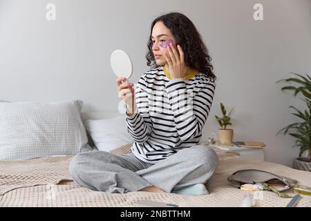 Porträt einer jungen, schönen, lockigen, dunkelhaarigen Frau, die lilafarbene Augenpflaster aufträgt und in einen kleinen Spiegel schaut, während sie auf dem Bett sitzt. Stockfoto