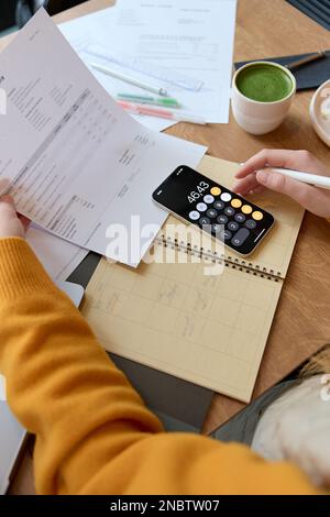 Draufsicht einer Unternehmerin, die am Tisch in einem Café sitzt, mit Notizen, die sich um persönliche Finanzen kümmern, und mit dem Taschenrechner auf dem Mobiltelefon Stockfoto