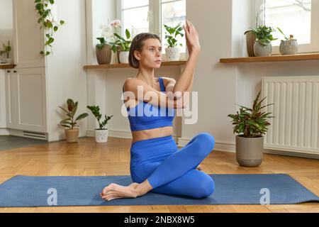 Porträt einer jungen, wunderschönen barfuß fokussierten Frau mit passendem Körper, die blaue Sportbekleidung trägt, in Gomukhasana sitzt und während des Yogas das Kuhgesicht auf der Yoga-Matte posiert Stockfoto