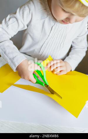 Süßes kleines Mädchen, das im Kindergarten Papierformen schneidet. Kind stellt Bewerbungen aus gelbem Papier Stockfoto