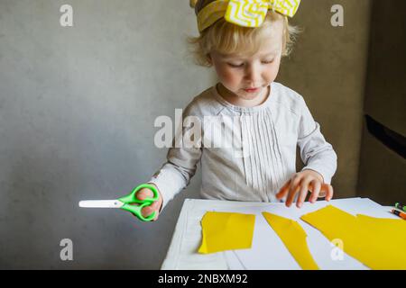 Kleines blondes Mädchen mit Schere in der Vorschule. Porträt eines kleinen süßen Mädchens, das eine Zeitung schneidet. Stockfoto