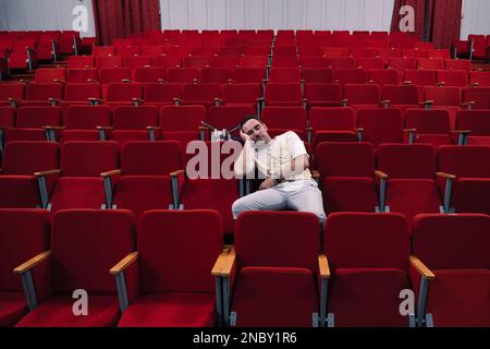 Junger Mann mit Brille schläft allein im Kinosaal, während alle weg sind. Ein einsamer Mann in einem leeren Theater schlief bei der Vorstellung ein. Ein Unint Stockfoto