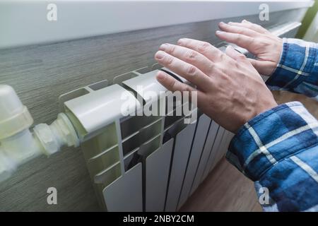 Nahaufnahme eines Mannes Hände, die sie über einer weißen Batterie erwärmen. Zum Begriff der Heizungsprobleme im Winter. Die Energiekrise. Stockfoto