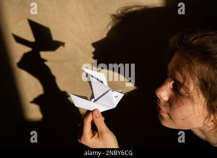 Kontrastierende Schatten an der Wand von einer Friedentaube aus Papier und dem Gesicht einer Frau. Stoppt den Krieg in der Ukraine. Spiel von Schatten und Licht. Dramatische Frau Stockfoto