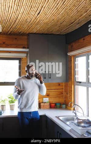 Ein afroamerikanischer Senior-Mann, der über ein Smartphone sprach, während er an der Küchentheke in der Blockhütte stand. Kopierraum, Technologie, Glatze, Brille, Unaltere Stockfoto