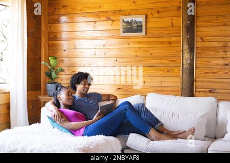 Glückliches afroamerikanisches Paar, das Zeit in einer Blockhütte mit einem Tablet verbringt. Blockhütte, Natur- und Lifestyle-Konzept. Stockfoto