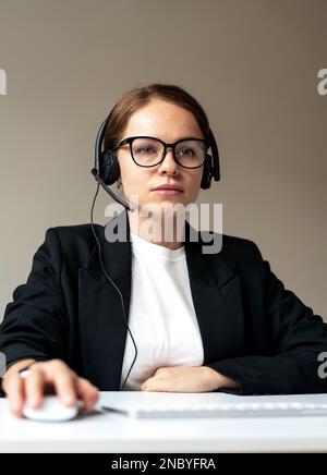 Aufnahme einer seriösen Büroangestellten mit Brille und Headset, die am Schreibtisch sitzt und am Computer arbeitet. Stockfoto