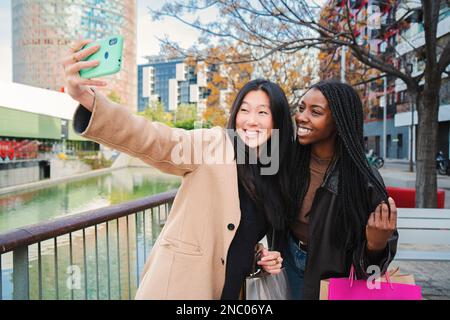 Ein paar multirassische glückliche Mädchen, die Spaß haben, ein Foto mit einem Handy zu machen. Zwei lächelnde junge Frauen, die an einem Einkaufstag ein Selfie-Porträt mit einem Smartphone machen. Lifestyle-Konzept. Hochwertiges Foto Stockfoto