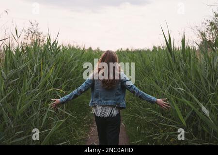 Ein fröhliches junges Mädchen mit langen braunen Haaren, das eine Jeansjacke trägt, mit offenen Armen und Händen, die das Gras berühren, auf der Wiese spazieren geht. Stockfoto