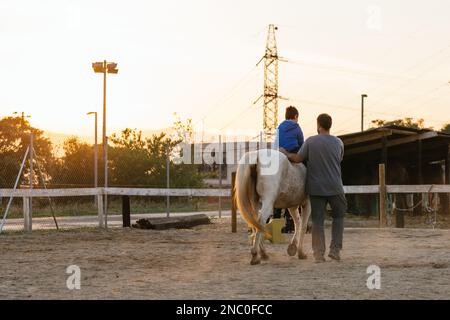 Ein Junge mit Behinderung reitet bei Sonnenuntergang im Reitzentrum. Stockfoto