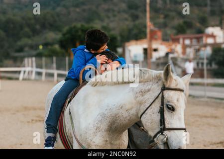 Ein behinderter Junge sitzt auf einem Pferd und umarmt seinen Pferdetherapielehrer. Stockfoto