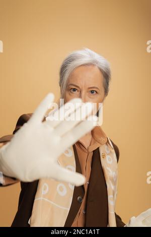 Seniorin mit weißem Handschuh, die in die Kamera schaut und ein Stoppschild auf beige zeigt Stockfoto