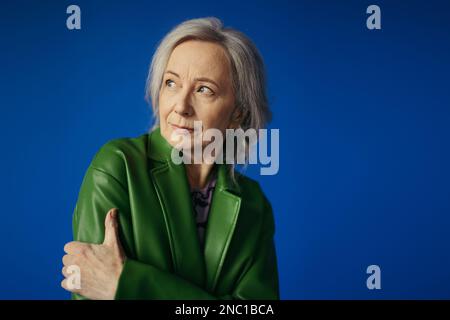 Eine pensive, grauhaarige Frau in grüner Lederjacke, die auf blau isoliert wegsieht Stockfoto