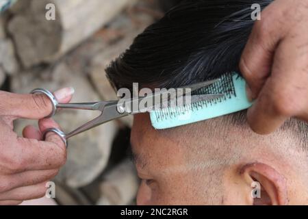 Tolle Zeit im Friseursalon. Haare schneiden, fröhlicher junger bärtiger Mann, der vom Friseur geschnitten wird und im Friseursalon auf einem Stuhl sitzt, Stockfoto