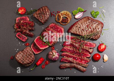 Gegrilltes Set mit verschiedenen Steaks mit Gemüse, Gewürzen und Kräutern. Klassische Fleischstücke umfassen Ribeye-, Augen-, Flanken- und Striploin-Steaks. Schwarzer Stein Stockfoto