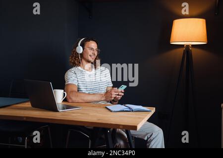 Ein junger, lächelnder Mann mit Kopfhörern, der ein Smartphone benutzt, während er an einem Laptop arbeitet, der zu Hause am Tisch sitzt Stockfoto