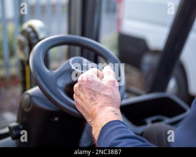 Ältere mitarbeiter nehmen das Lenkrad eines Gabelstaplers am Arbeitsplatz in die Hand und nehmen nach 70 Jahren wieder an der Arbeit Teil Stockfoto