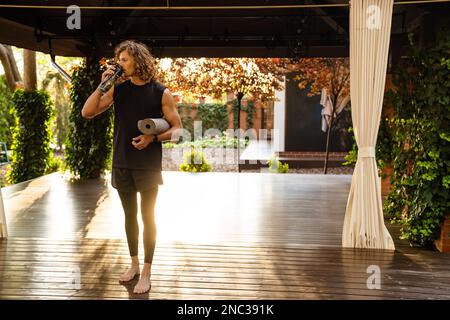 Junger, lockiger Mann mit Trainingsmatte und Trinkwasser im Yoga-Studio im Freien Stockfoto