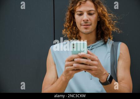 Porträt eines jungen, gutaussehenden, langhaarigen Sportlers, der sein Handy benutzt, während er an der grauen Wand draußen steht Stockfoto