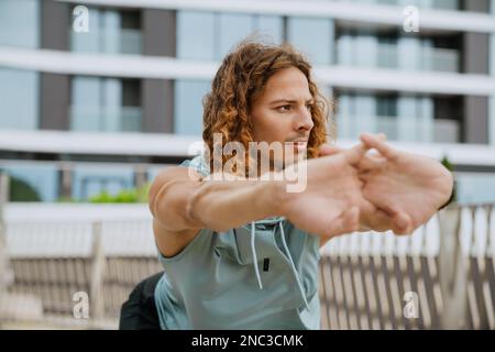 Ein junger, sportlicher, langhaariger, gutaussehender Mann, der sich zur Seite streckt, während er draußen steht Stockfoto