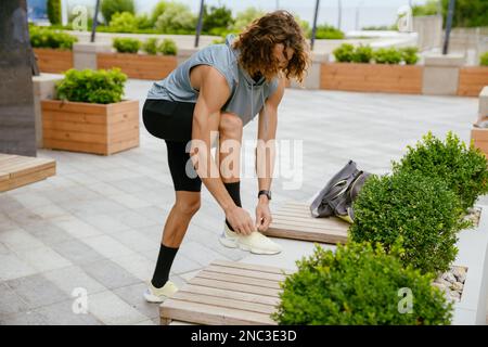 Porträt eines jungen, langhaarigen Sportlers, der im Freien Schnürsenkel bindet und ein Bein auf einer Bank legt Stockfoto