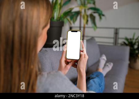 Rückansicht eine unbekannte, lässig blonde junge Frau in einem modernen Wohnzimmer, die auf einem Sofa mit einem iPhone-Smartphone-Modell und einem leeren Bildschirm arbeitet Stockfoto
