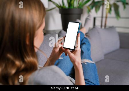 Rückansicht eine unbekannte, lässig blonde junge Frau in einem modernen Wohnzimmer, die auf einem Sofa mit einem iPhone-Smartphone-Modell und einem leeren Bildschirm arbeitet Stockfoto