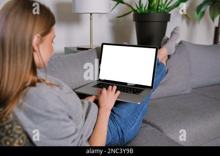 Nicht wiedererkennbare, lässig blonde junge Frau in einem modernen Wohnzimmer, die auf einem Sofa liegt und mit überkreuzten Beinen arbeitet, mit einem Laptop-Modell mit leerem Bildschirm Stockfoto