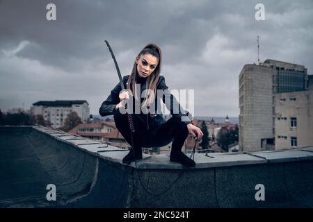 Eine coole weibliche Ninja-Kriegerin in Lederbekleidung, die ein japanisches Schwert auf einem Gebäude schwingt Stockfoto