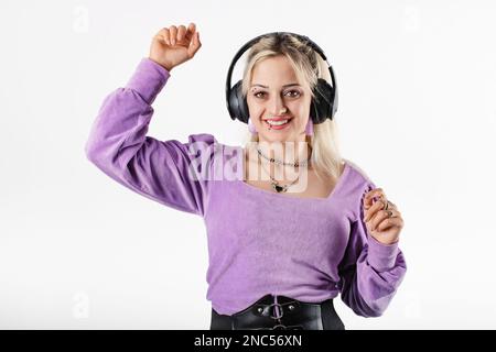 Die fröhliche blonde Frau trägt eine lila gerippte Bluse, isoliert auf weißem Hintergrund, lächelt in die Kamera und tanzt über Kopfhörer zur Musik. Stockfoto
