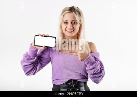 Das Porträt einer süßen blonden Frau, die isoliert auf weißem Hintergrund steht, das Telefon horizontal in der Hand hält, zeigt auf den leeren weißen Modellbildschirm mit Stockfoto