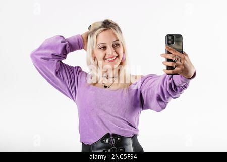 Wunderschöne blonde Frau mit gerippter Bluse, die isoliert auf weißem Hintergrund steht und das Haar berührt, während sie Selfie mit dem Mobiltelefon macht. Aufnahme von Fotos f Stockfoto