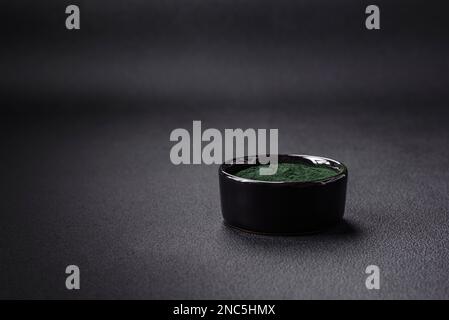 Gewürz, grüne Spirulina in Form von Pulver in einer schwarzen Schüssel auf dunklem Betonhintergrund Stockfoto