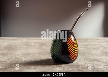 Hübscher, glänzender Keramik-Yerba-Mate-Becher mit orangefarbenem und grünem Blattdesign und Metallbombilla-Stroh auf einem Betontisch Stockfoto