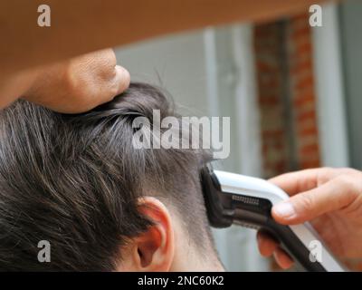 Zu Hause schneidet ein Mann mit einem elektrischen Trimmer lange Haare vom Hinterkopf des Mannes. Ein Mann schneidet einen jungen Mann mit einem elektrischen Rasierer von der Seite Stockfoto