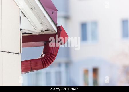 Ein Schneetropfen auf der roten Dachrinne eines Gebäudes vor dem Hintergrund eines Hauses. Foto in hoher Qualität. Horizontal. Stockfoto