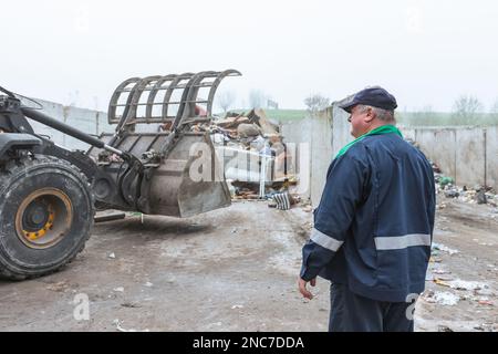 Ein Arbeiter im Recycling-Center, in dunkelblauer Arbeitskleidung, der sich einen unsortierten Müllhaufen ansieht, ist gerade in der Materialrückgewinnungsanlage angekommen, Rückansicht. Stockfoto
