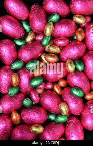 Stapel oder Gruppe von bunten, in Folie verpackten Schokoladeneiern in verschiedenen Größen in Pink, Rot, Gold, Orange und Limettengrün. Stockfoto