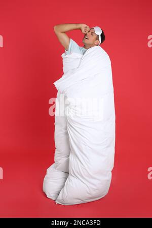 Mann mit Schlafmaske, eingewickelt mit Decke auf rotem Hintergrund Stockfoto