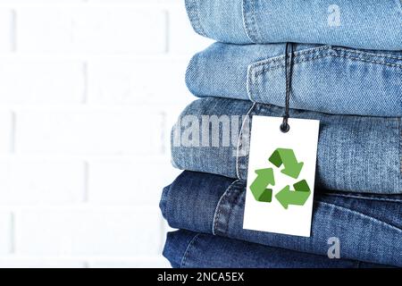 Ein Stapel verschiedener Jeans mit Recycling-Etikett in der Nähe von weißen Backsteinwänden, Nahaufnahme. Platz für Text Stockfoto