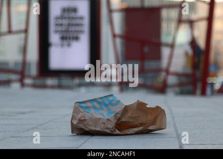 Fast Food Papiertüte als Müll auf der Straße mit einer Bushaltestelle Hintergrund. Gebrauchte Beutel aus Lebensmitteln, die im Fastfood-Restaurant gekauft wurden Umweltverschmutzung Stockfoto