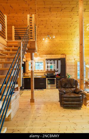 Treppe mit Holzpfosten und schwarzem Metallrohr-Handlauf plus braunem Ledersessel und Küchenbereich im Hintergrund im Blockhaus. Stockfoto