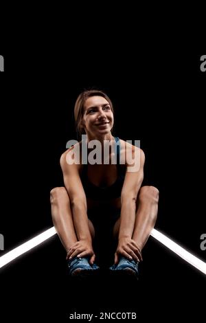 Weibliche Läuferin, die auf dem Boden ruht. Mädchen in Sportbekleidung, die auf einer beleuchteten Rennstrecke vor dunklem Hintergrund sitzt. Stockfoto