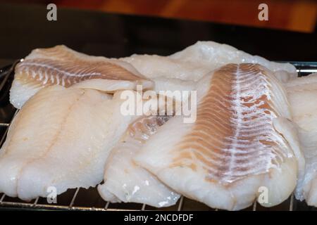 Nahaufnahme mit mehreren Stücken rohen dicken, gesunden weißen Fischen ungekochtes Kabeljaufilet oder Kabeljaufilets, die auf einem Drahtgestell mit grauem Hintergrund getrocknet werden. Stockfoto