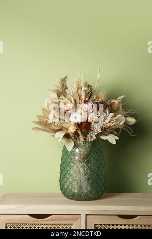 Wunderschöner getrockneter Blumenstrauß in Glasvase auf einem Holztisch nahe der Olivenwand Stockfoto