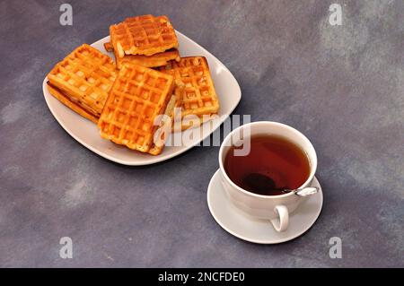 Ein Teller mit Wiener Waffeln und eine Tasse heißem schwarzen Tee auf grauem abstraktem Hintergrund. Nahaufnahme. Stockfoto