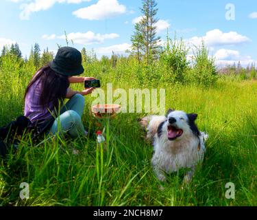 Ein Mädchen mit Hut, das auf dem grünen Gras sitzt, macht ein Selfie am Telefon mit einem weißen, fröhlichen Hund im Sommer im Wald von Yakutia in Sibirien. Stockfoto