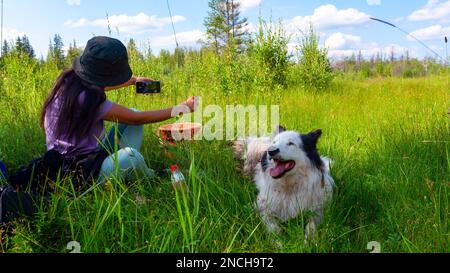 Ein Mädchen mit Hut, das auf dem grünen Gras sitzt, macht tagsüber ein Selfie am Telefon mit einem weißen, fröhlichen Hund im Wald. Stockfoto